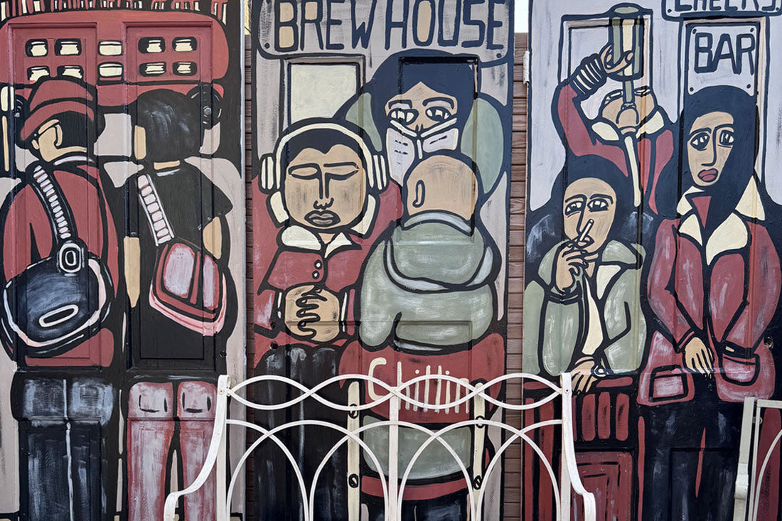 Hand-painted garden mural design by ATADesigns, featuring bold colourful figures in a street bar and coffee shop scene, installed in a garden with a garden chair in front.
