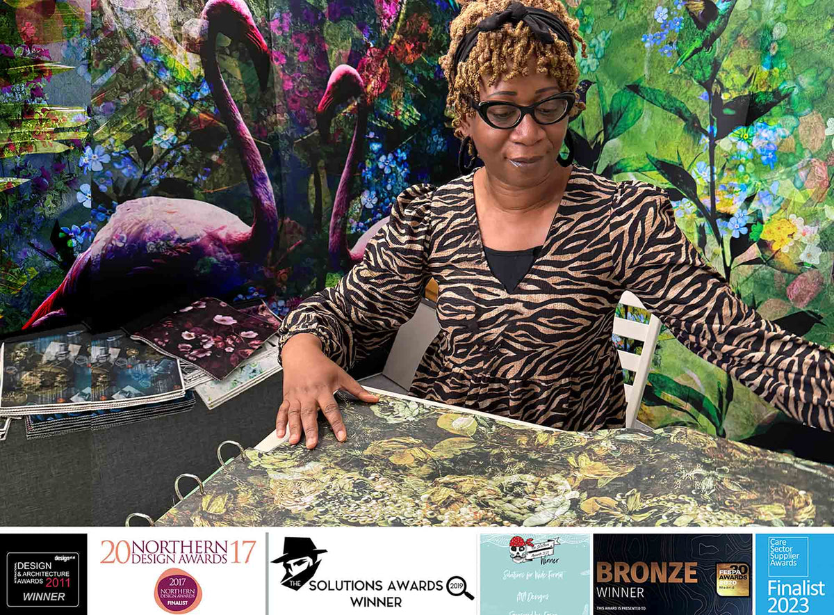 Annette Taylor-Anderson sitting at a table with artwork, surrounded by colorful flamingo mural wallcovering.  Mural design by ATADesigns.