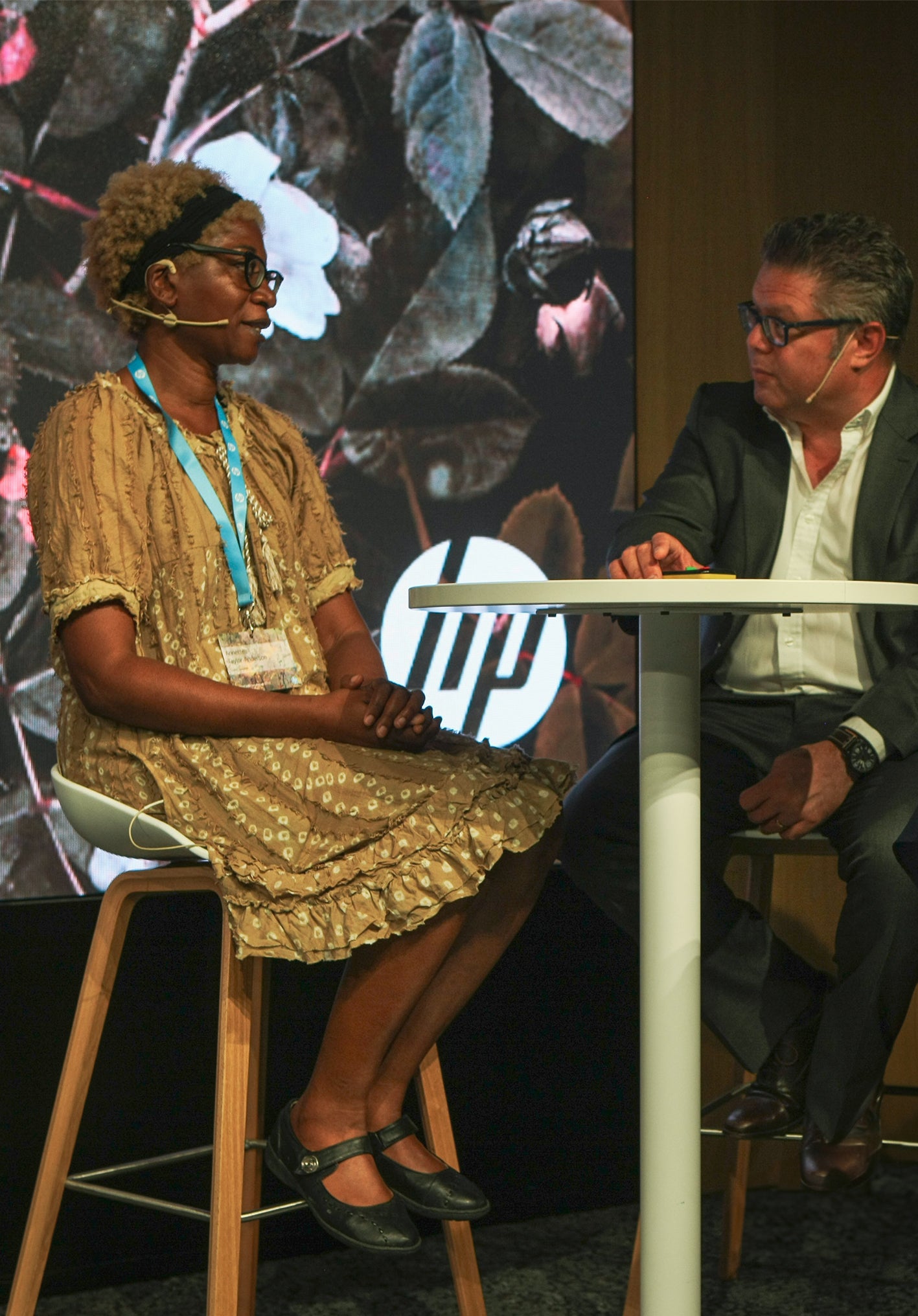 Annette Taylor-Anderson and erry Raghunath sitting at a table in front of mural with HP logo wearing microphones, talking.