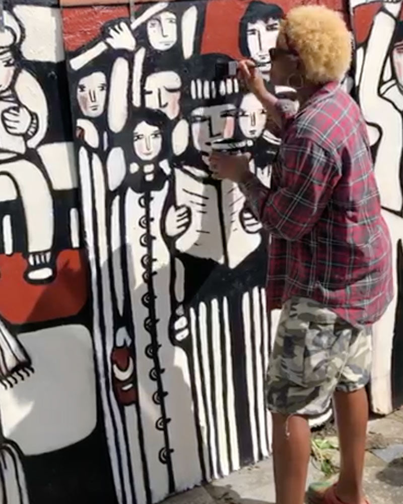 A person with blonde hair stands beside a large painted mural, holding a paintbrush and adding to the artwork. The mural features bold black, white, and red abstract figures and faces.