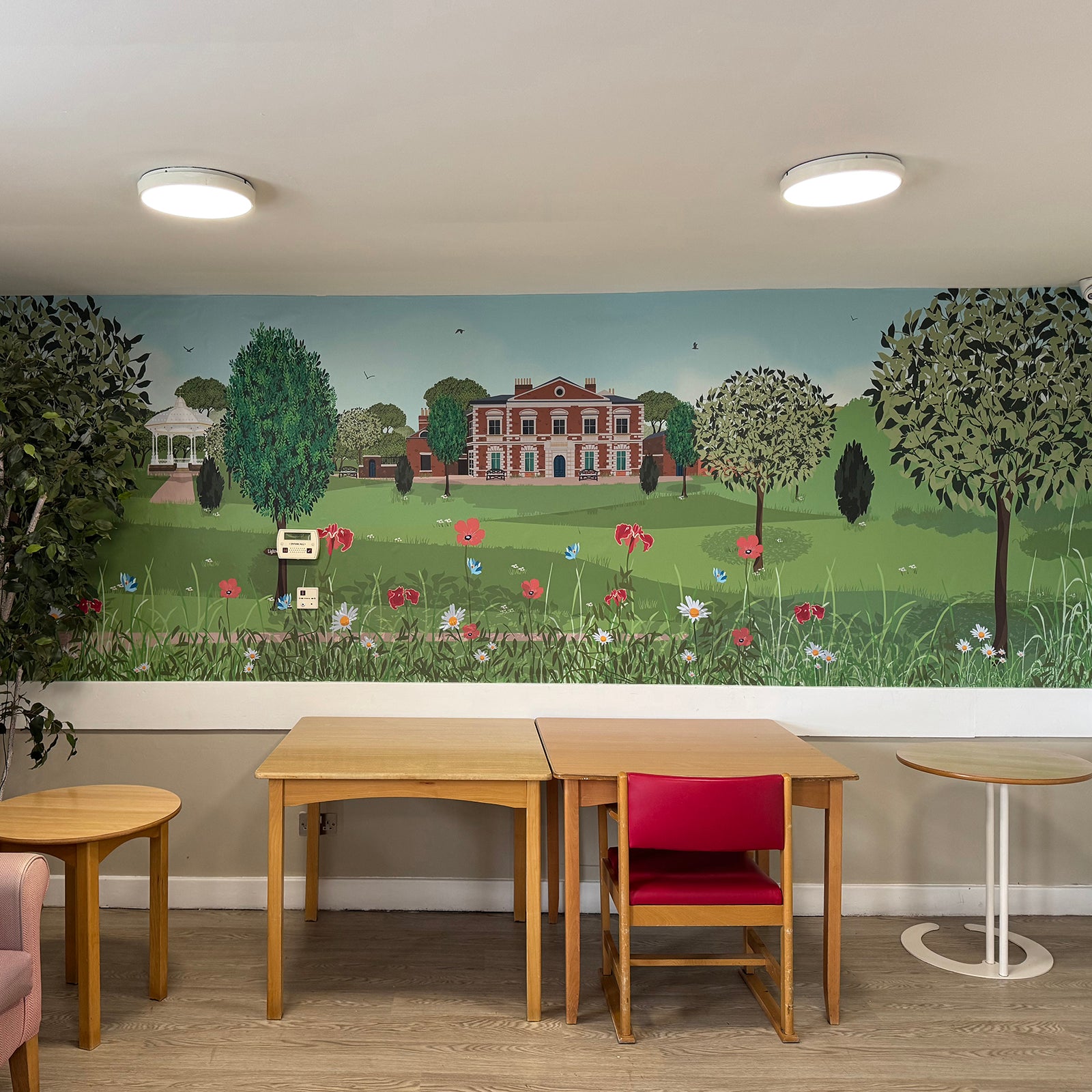 Newbury Manor Nursing Home with tables and red chair in front of mural illustration design by ATADesigns.  The mural features Lightwoods Park and House, flowers in foreground with trees a bandstand in the background.