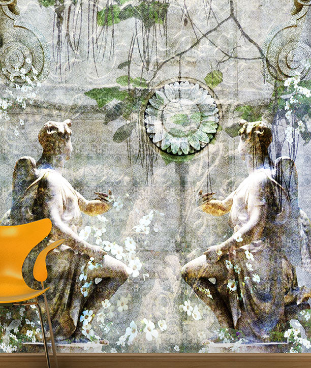 A wall mural Angels Eden featuring two classical winged female statues seated in a garden scene with hanging vines and a circular floral medallion, shown installed on a wall above a wooden baseboard. A small orange chair is partially visible in the lower left corner. Mural design by ATADesigns.