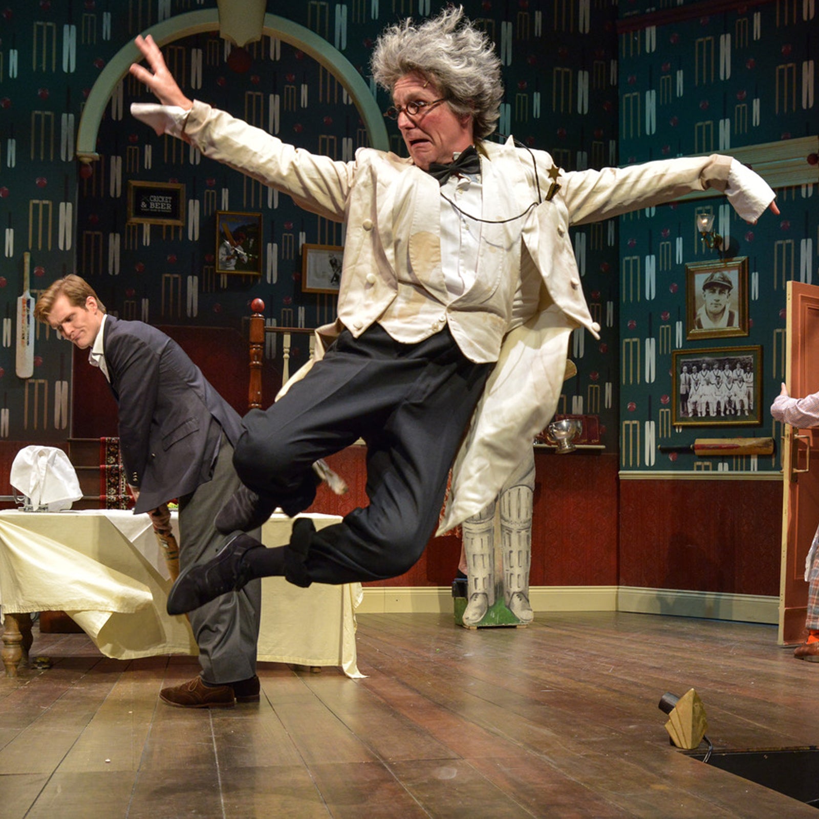 Person in a theatrical setting with a dynamic pose, wearing a white suit and grey hair.   Also featuring a man in a grey suit behind playing cricket.  Cricket mural in the background designed by ATADesigns.