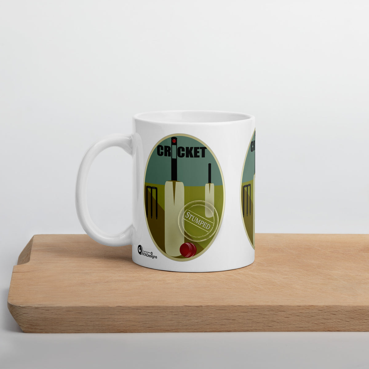  White Cricket mug with cream bats, black stumps and a red ball on a teal and olive background, displayed on a wooden board.