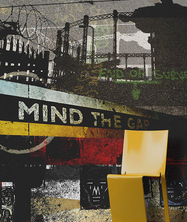A modern yellow chair positioned in front of a highly textured, grunge-style collage wall mural featuring the phrase 'MIND THE GAP' and industrial imagery. Mural design by ATADesigns.