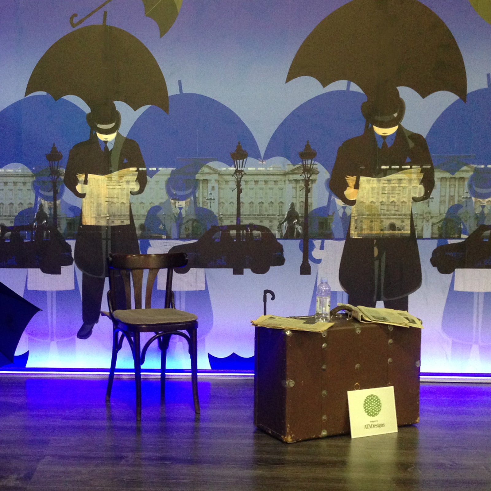 Neon Palace mural design by ATADesigns featuring large scale gentlemen wearing bowler had long coat, shirt and tie, holding black umbrella reading a newspaper with Buckingham Palace and black taxis in the background.  The mural is featured as a backdrop in an exhibition space with a single chair stool and large suitcase on the floor.