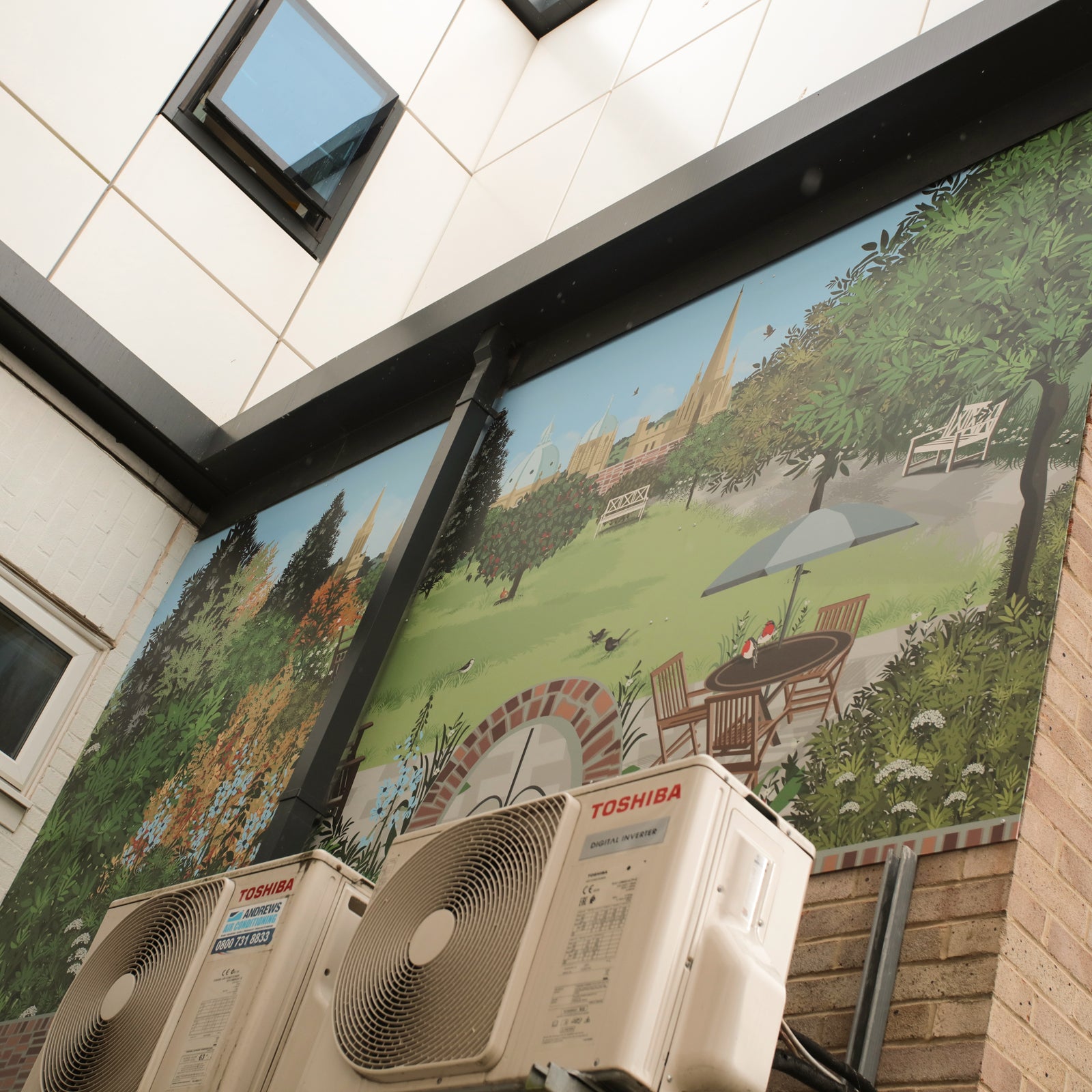 Outdoor garden mural design featuring the city of oxford in the background. Mural design by ATADesigns.  Installed building on brick wall high up near windows of white building.