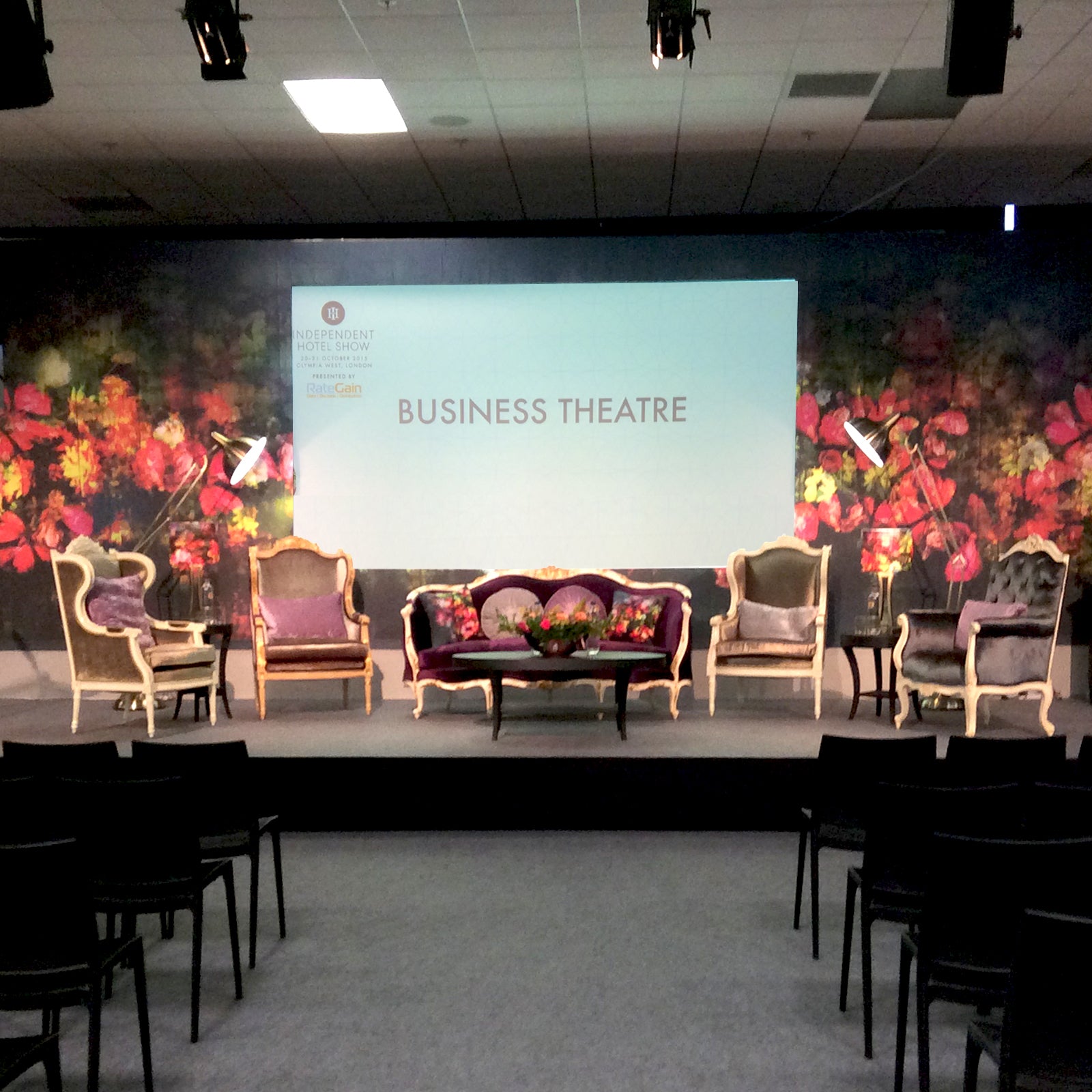 Business theatre setting with screen and Regents Floral  Expression colourful mural design backdrop by ATADesigns.  Armchairs on stage with table, with audience chairs facing screen.