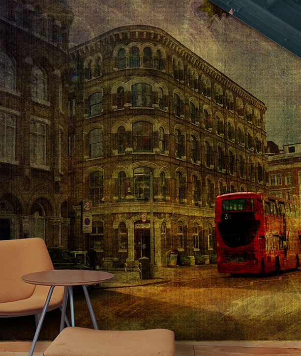 A vintage-style wall mural depicting a London Southwark street scene with a red double-decker bus and old brick buildings, shown installed in an interior setting with a small round table and chair in the foreground. Mural design by ATADesigns.
