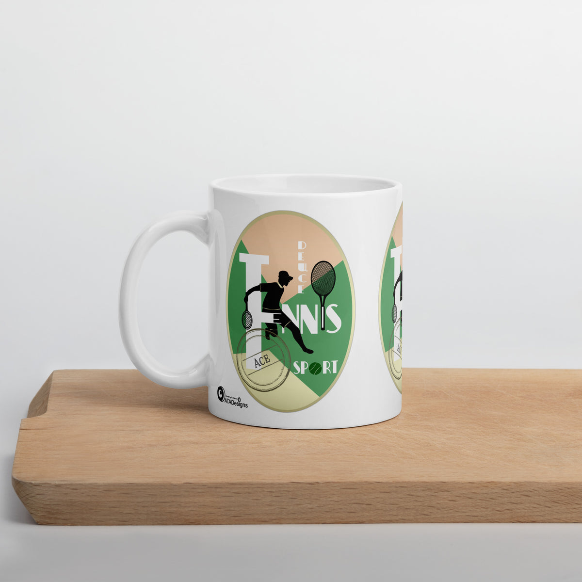 White Tennis Deuce mug with a black tennis-player silhouette and green geometric artwork, displayed on a wooden board.