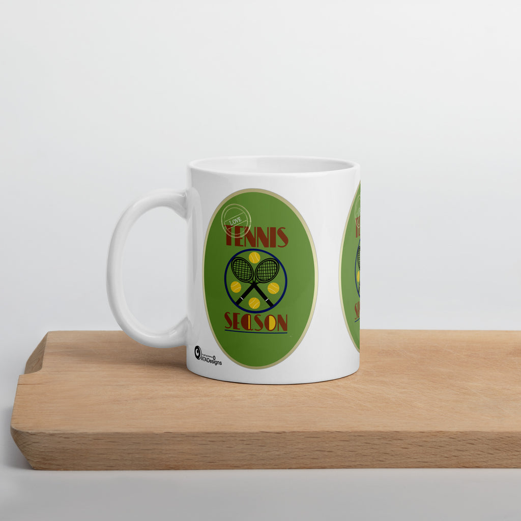 White Tennis Season mug with crossed rackets, yellow tennis balls and green artwork, displayed on a wooden board.